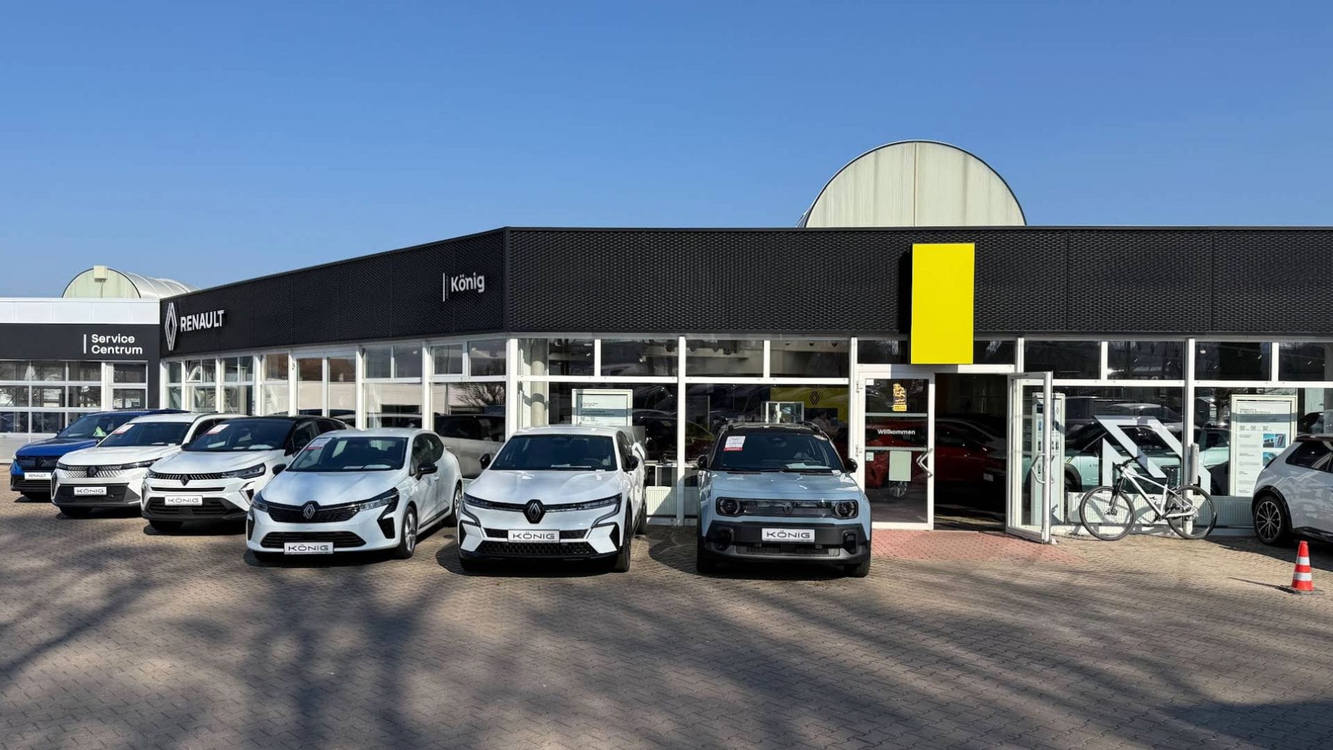 Frontansicht eines Renault Autohauses von König mit Glasfassade und Servicebereich, davor stehen mehrere Fahrzeuge in einer Reihe. Der Eingangsbereich ist geöffnet, daneben ist ein Fahrrad abgestellt, das Renault-Logo ist deutlich sichtbar.