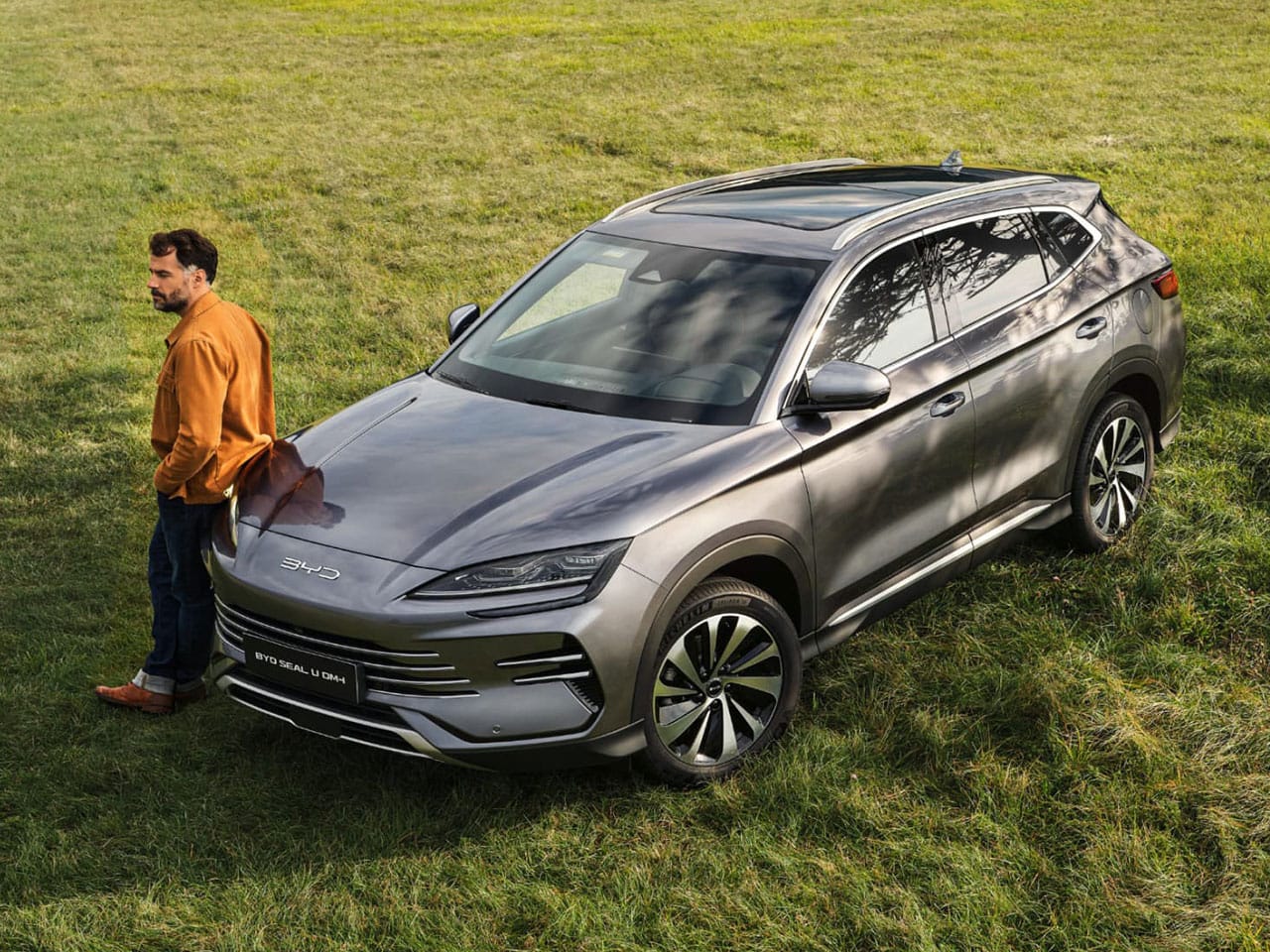 Silbernes Auto BYD Seal U steht auf einer Wiese und ein Mann in orangener Jacke lehnt entspannt am Auto