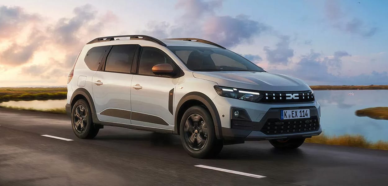 Ein hellgrauer Dacia Jogger fährt auf einer Straße durch eine schöne Seen-Landschaft.
