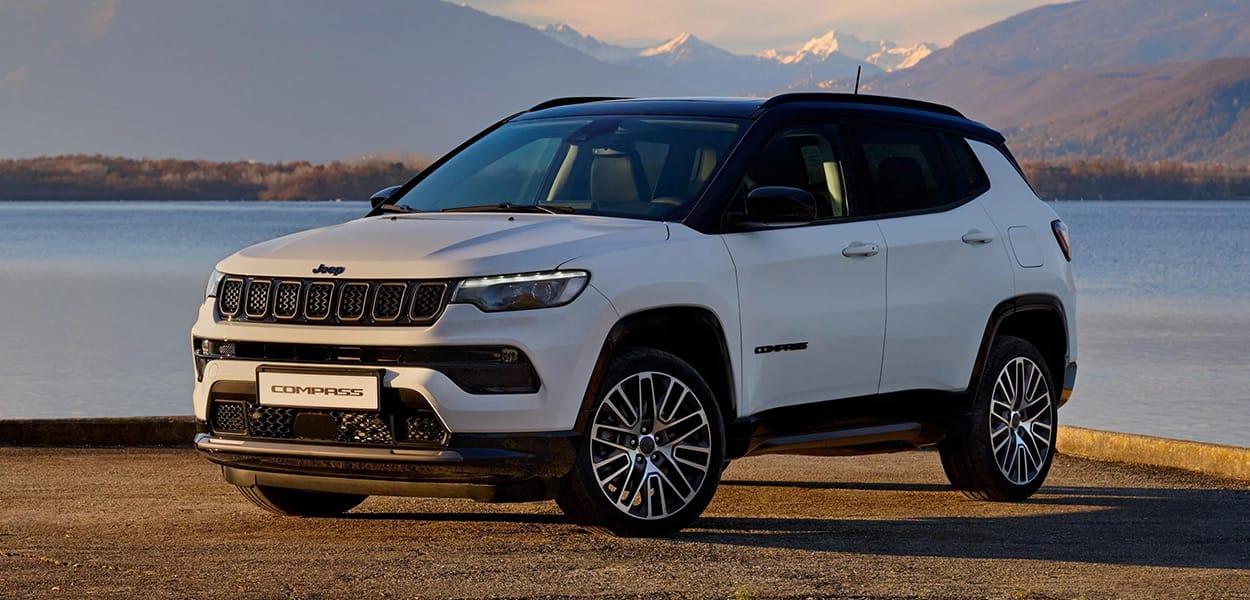 Ein weiß-schwarzer Jeep Compass steht auf einem Platz an einem See vor einer Berg-Kulisse.