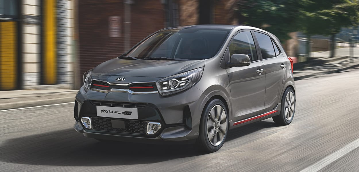 Ein grausilberner Kia Picanto mit roten Streifen fährt durch eine Stadt eine Straße entlang.