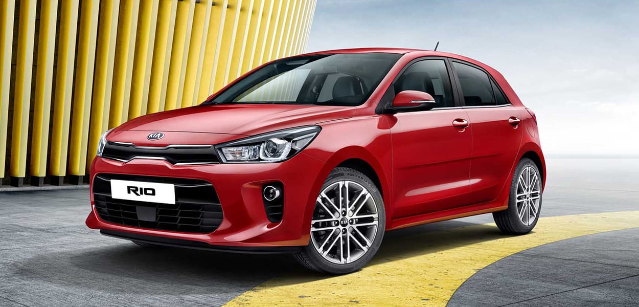 Ein roter Kia Rio steht auf einem Platz vor einem modernen gelben Gebäude.
