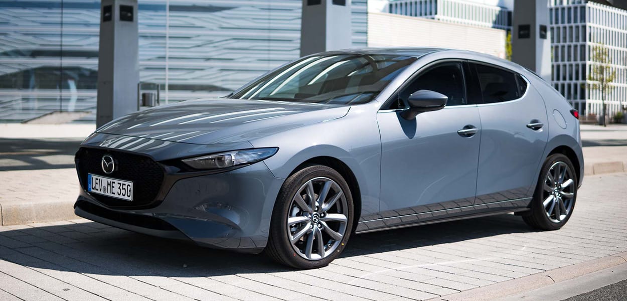 Ein blaugrauer Mazda 3 steht auf einem Parkplatz vor einer modernen Stadtkulisse.