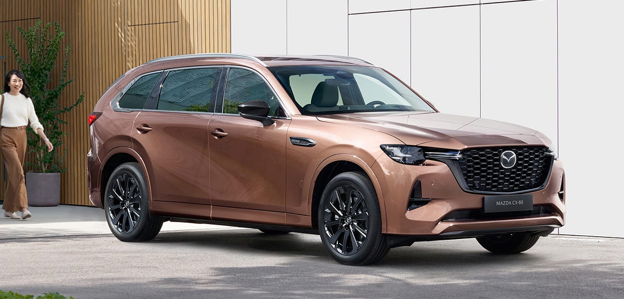 Ein hellbrauner Mazda CX-80 steht auf einem Parkplatz vor einem Gebäude.