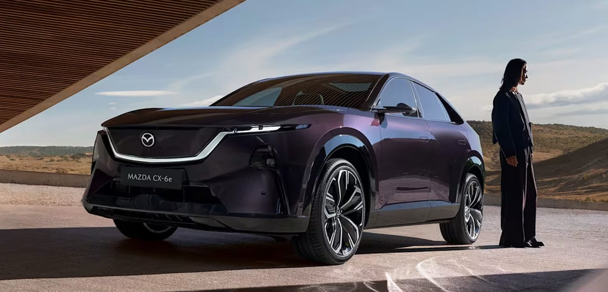 Ein violetter Mazda CX-6e steht mit einer Person auf einem Platz an einem modernen Gebäude.