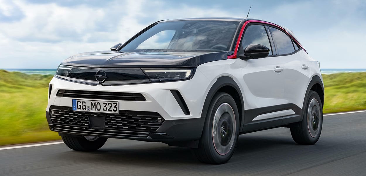 Ein weißer Opel Mokka mit schwarzer Motorhaube und rotem Zierstreifen fährt über eine Landstraße.