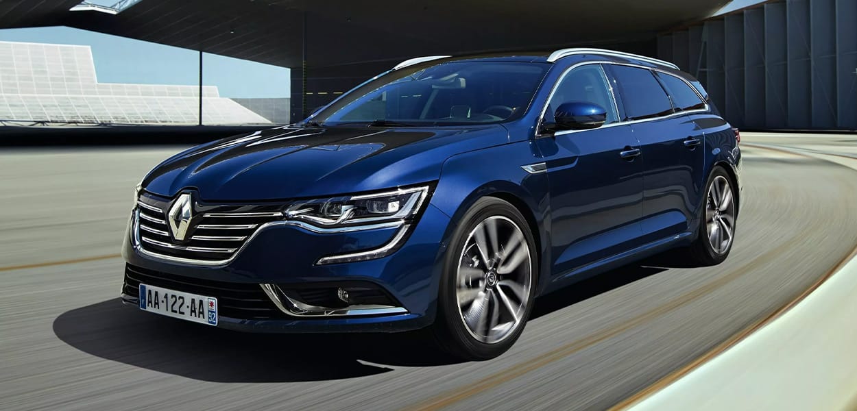 Ein dunkelblauer Renault Talisman Kombi fährt um eine Kurve an einem modernen Hangar vorbei.