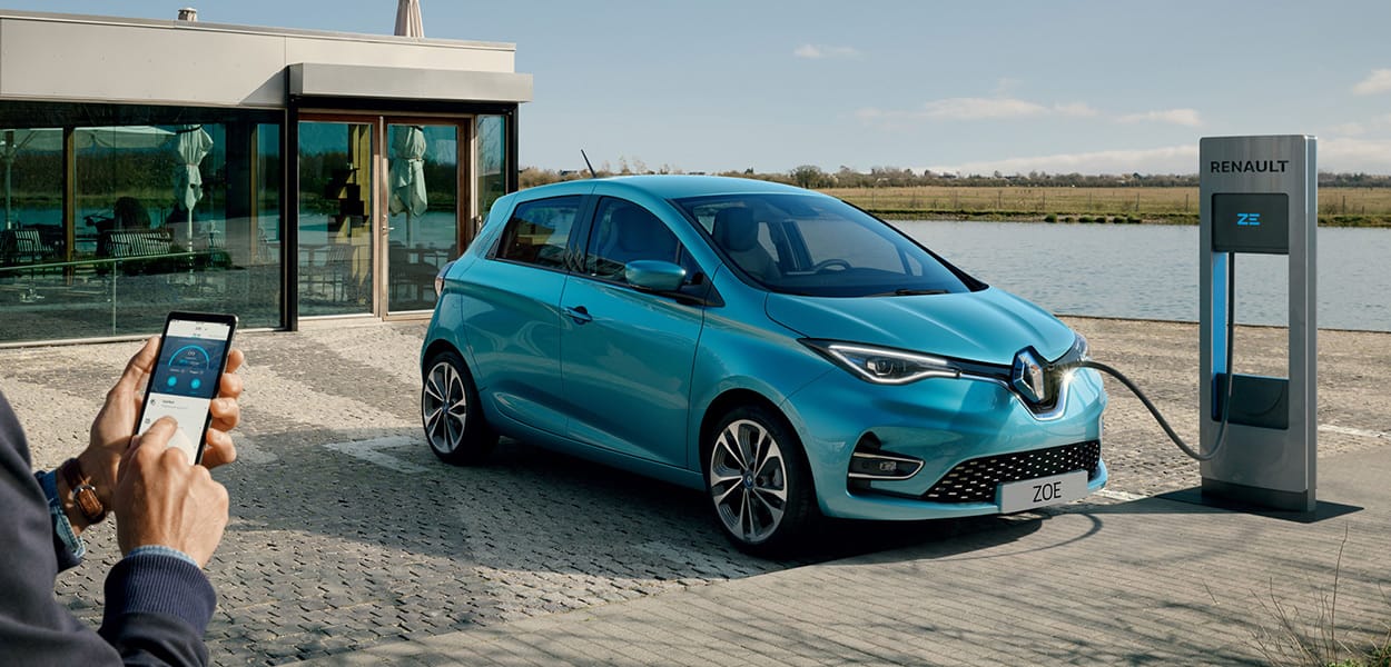 Ein blauer Renault Zoe lädt an einer Ladesäule am Wasser vor einem Gebäude, während das Smartphone mit einer Lade-App von zwei Händen bedient wird.