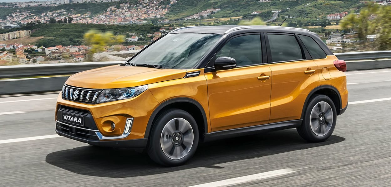 Ein orangener Suzuki Vitara fährt eine Landstraße mit Leitplanke entlang an einer bergigen Landschaft vorbei.